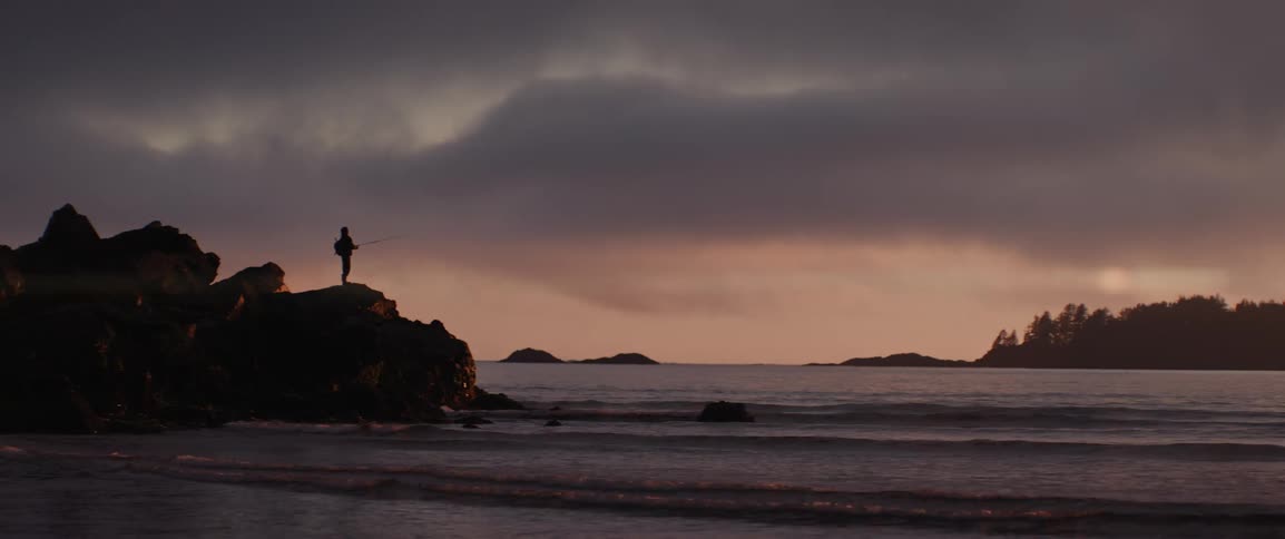 Person standing on a rock near the beach against a red horizon casts a fishing line