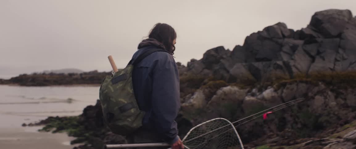 Man carries a fishing rod and net onto a rocky outcrop on the beach