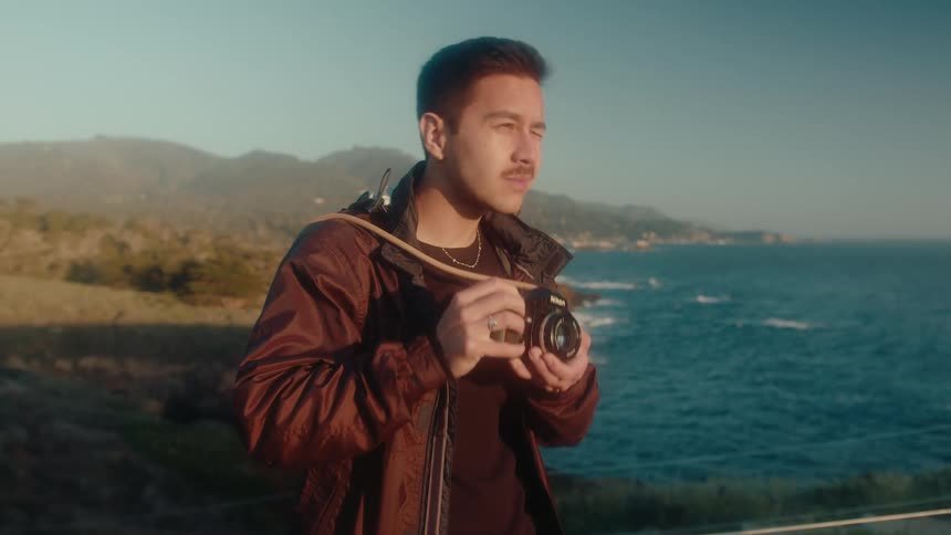 A photographer with a mustachea black shirt and a crimson jacket takes photos of the coast with a camera