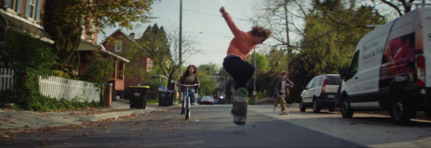 Three children are playing on a suburban street riding skateboards and bicycles