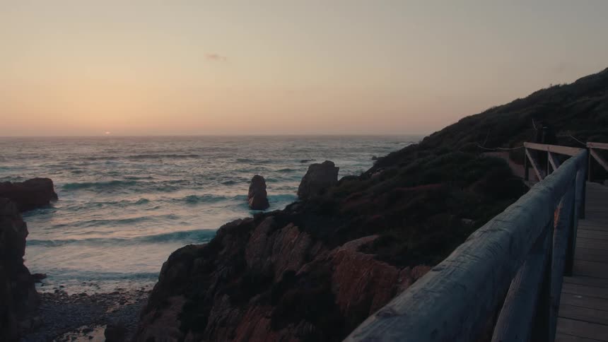 A wooden walkway overlooking a rocky coastline at sunset with waves crashing against the cliffs