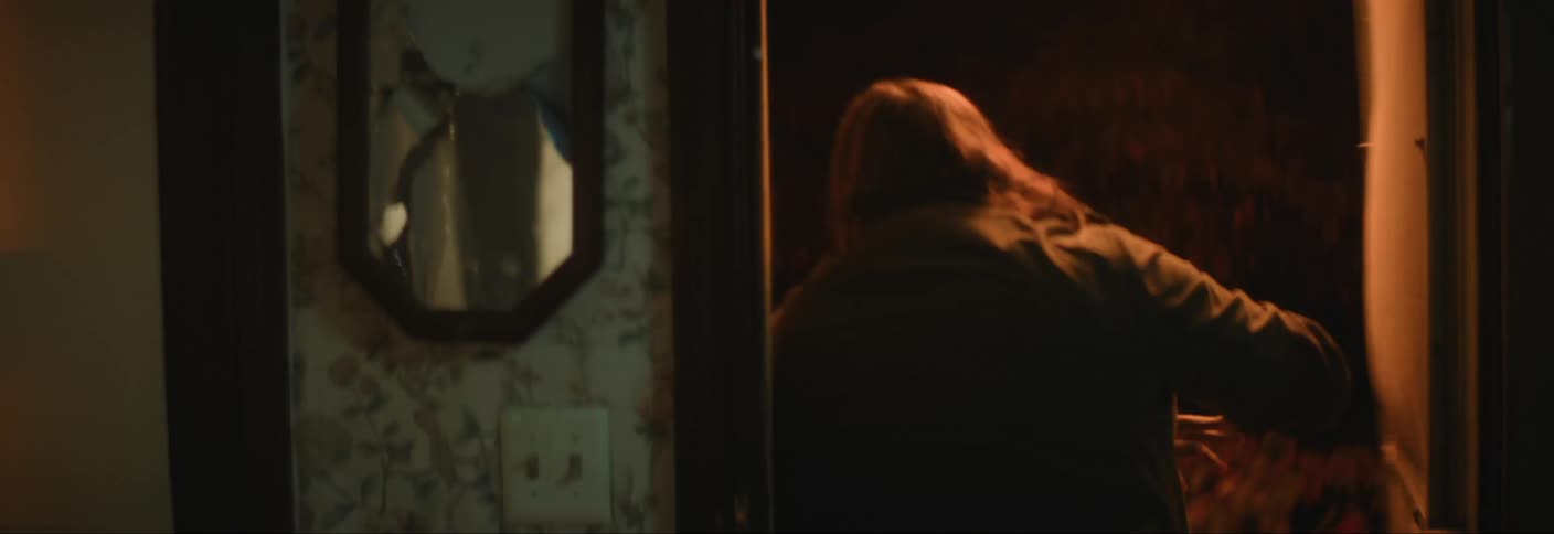 A person shatters a mirror on the wall in a dimly lit room and exits the door in anger