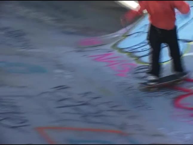 A person wearing a red sweatshirt and black pants is shown skateboarding in a graffiti covered skatepark