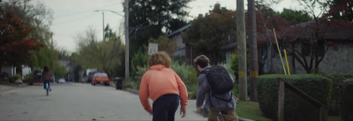 Siblings skateboard down a residential street while a woman cycles ahead of them in the distance on an autumn day