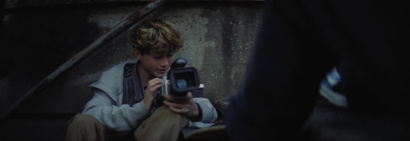 A young man in a grey jacket is holding a large camera as he speaks to someone