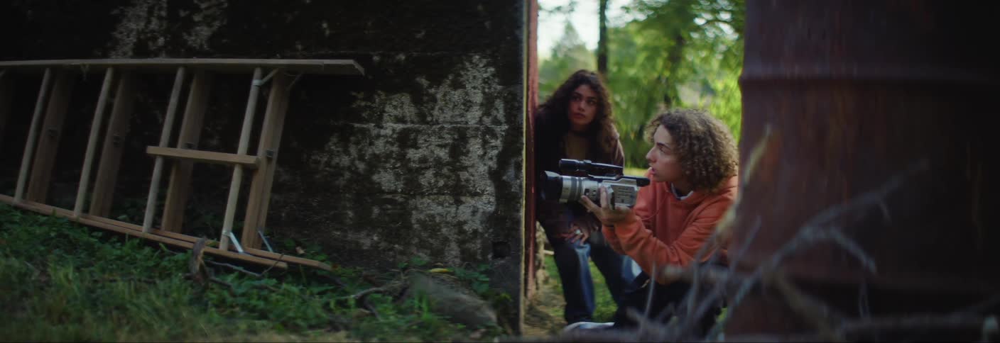 A group of kids filming with a camera hide behind a wall before running away in fear