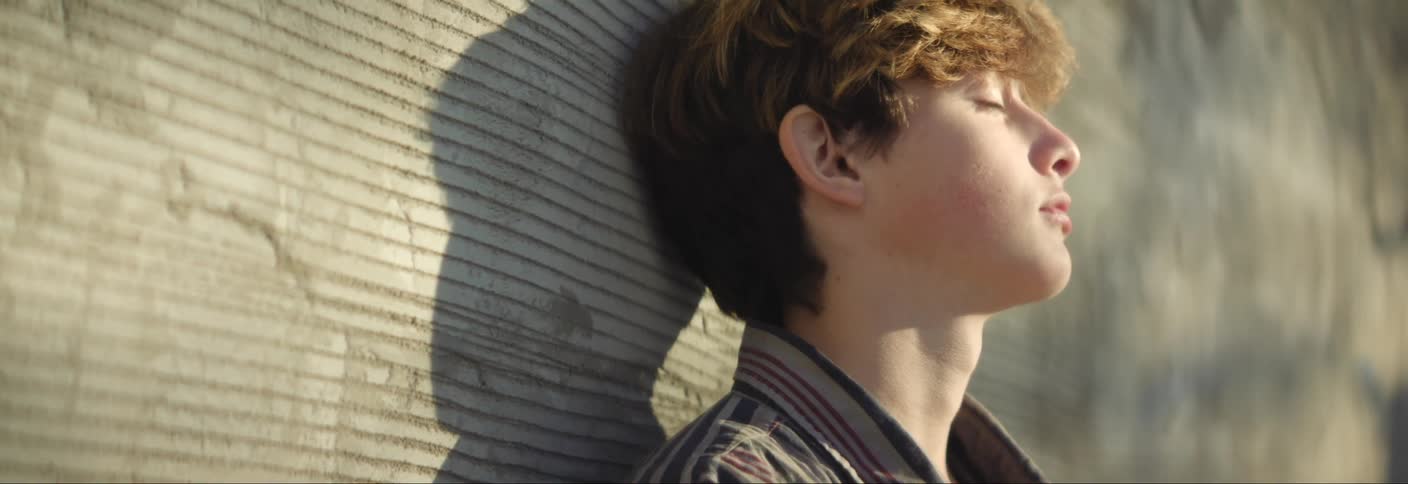 Teenager leaning against a wall with his eyes closed wipes his nose