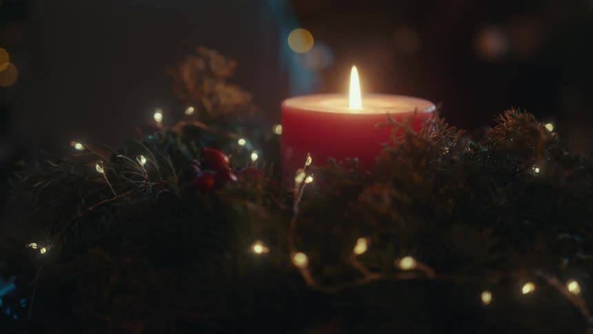 A lit red candle sits in the center of a festive arrangement of greenery and warm twinkling lights