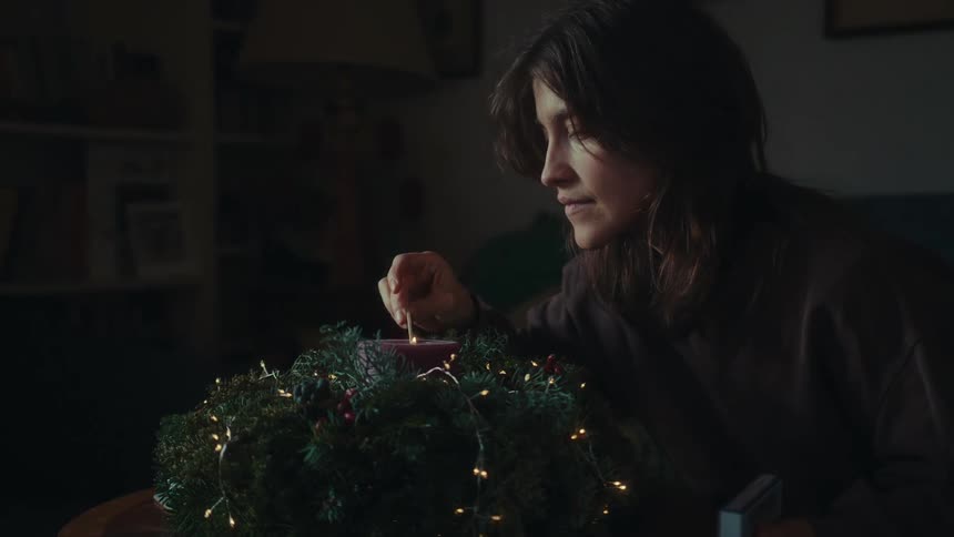 A solitary woman lights up a christmas candle on a dimly lit living room