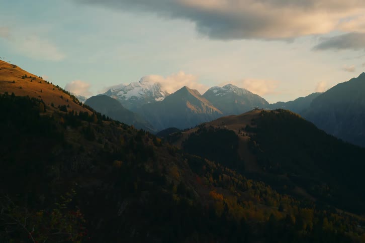 A landscape view reveals a series of mountain peaks some snow capped silhouetted against a sky with scattered clouds at dusk