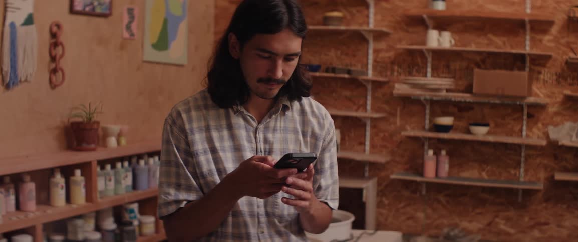 A person wearing a plaid shirt is looking at a smartphone while standing in a room with wooden shelves