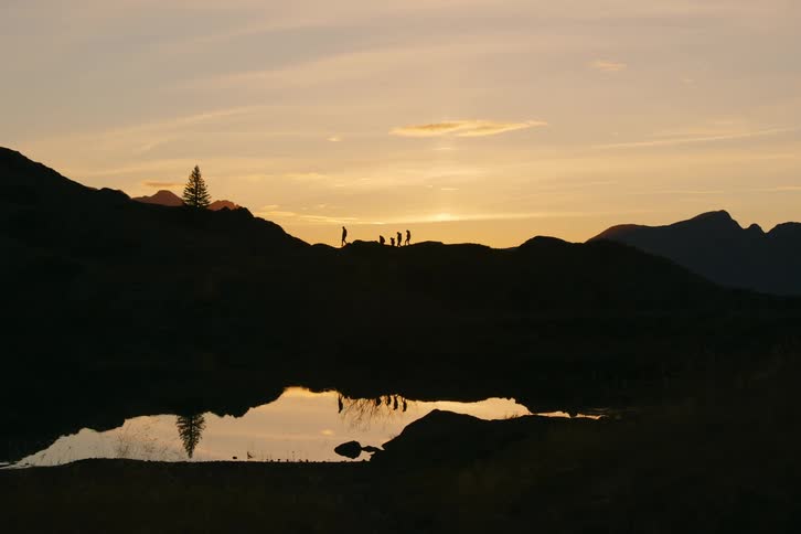 The silhouette of some people walking on top of a hill on a dark morning and their images are reflecting in a lake