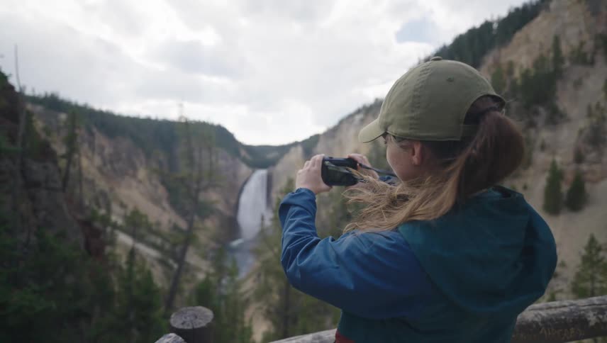 Woman stands near the rail at a lookout point and takes a picture of a waterfall on an overcast day