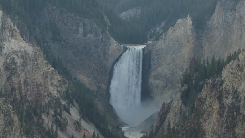 Waterfall flows down a cliff in the mountains into a river passing between slopes on a foggy day