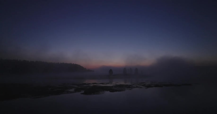 A still body of water reflecting the sky and a few silhouettes shrouded in a thick fog