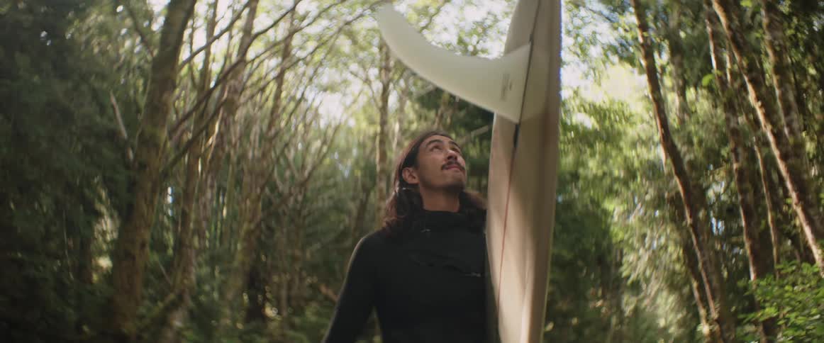 A clip of a man walking with a surfboard down a wooded path