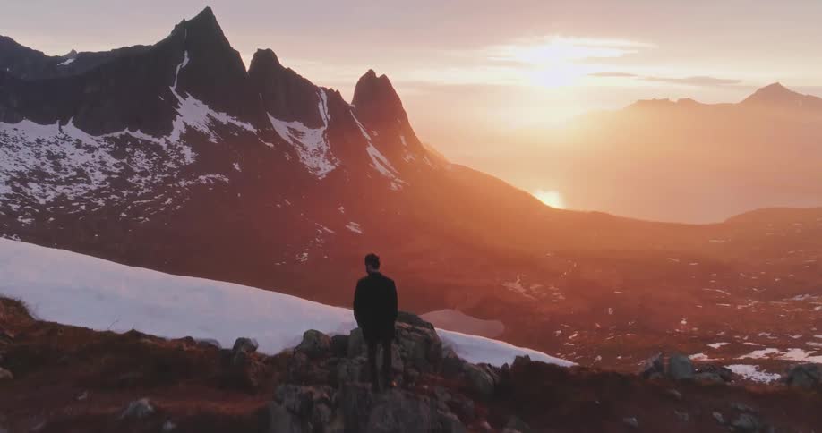 Man standing on a mountain overlooking a snow covered valley as the sun sets over mountains in the distance