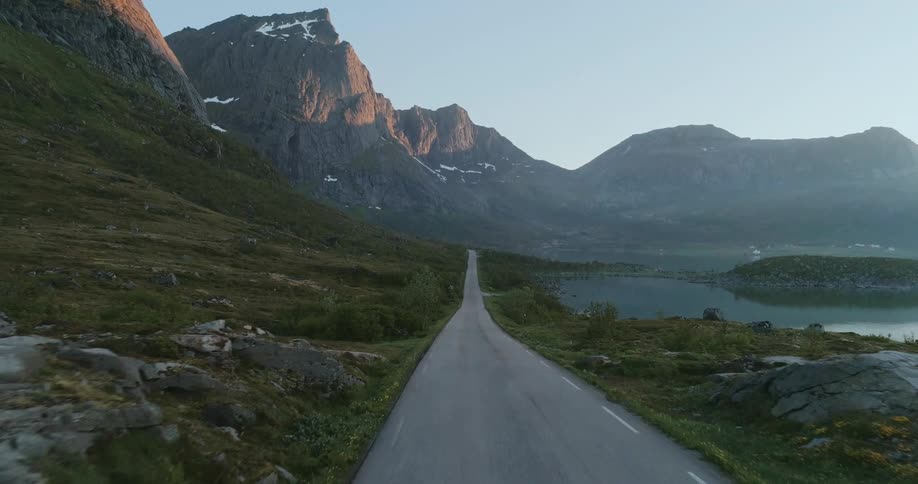 A winding asphalt road traverses a rugged landscape of green slopes and glimpses of a shimmering lake leading towards a panorama of distant mountains