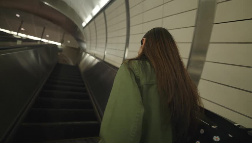 A woman in a green jacket talking on the phone while standing near an escalator in a welllit modern subway station