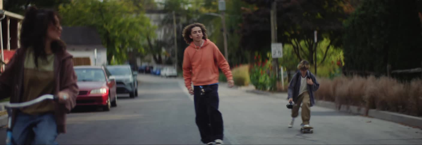 A teenage girl riding on an asphalt street on a bicycle and two boys on a skateboard and one of them is wearing an orange jacket and doing a trick