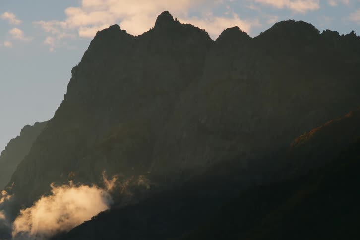 A mountain range silhouetted against a sunset sky with clouds casting shadows on the peaks