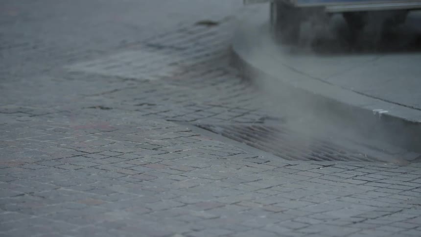 Steam is rising from a street drain next to a curb on a cobblestone road