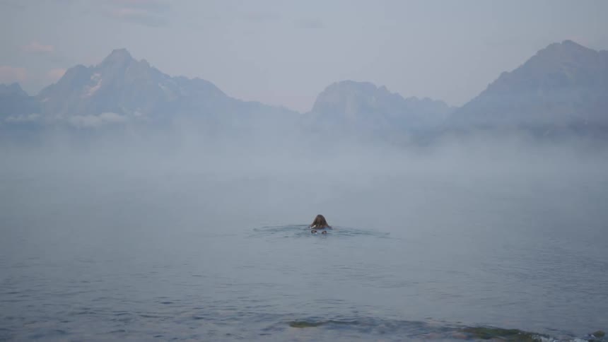 A woman from behind is entering a lake with thick fog and swimming forward