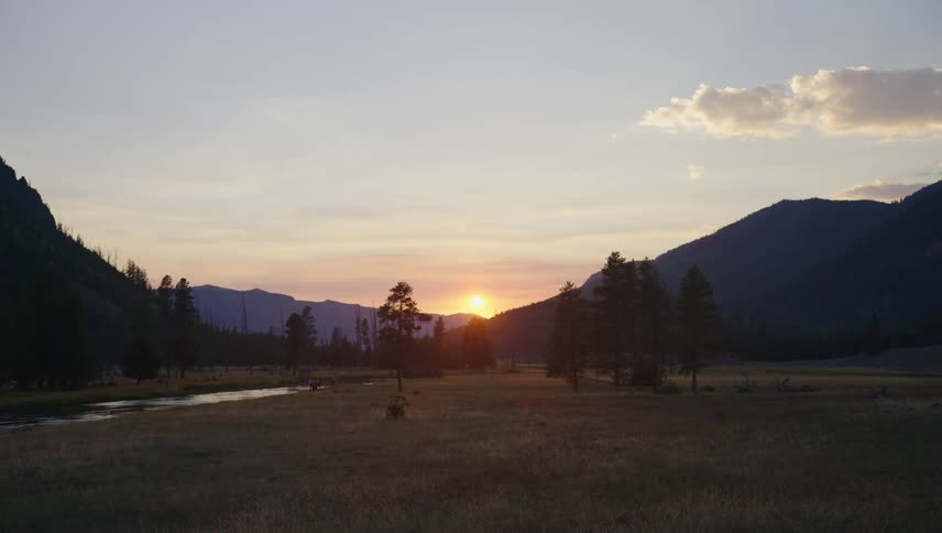 The sun sets behind mountains silhouetting trees against the bright sky while a group of people gather near a river in the distance