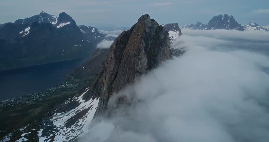 A mountain peak shrouded in mist with a coastal village and a fjord visible in the distance