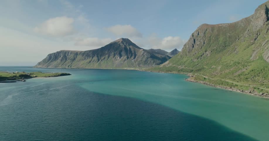 A calm bay with clear blue water is surrounded by steep rocky mountains with lush green slopes under a bright blue sky