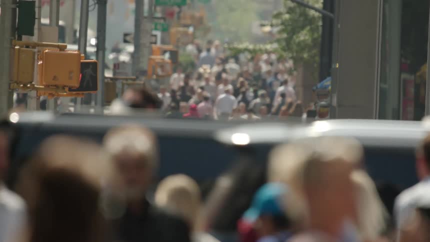 A street with a huge crowd of pedestrians passing by and a car passing on the street next to a traffic light