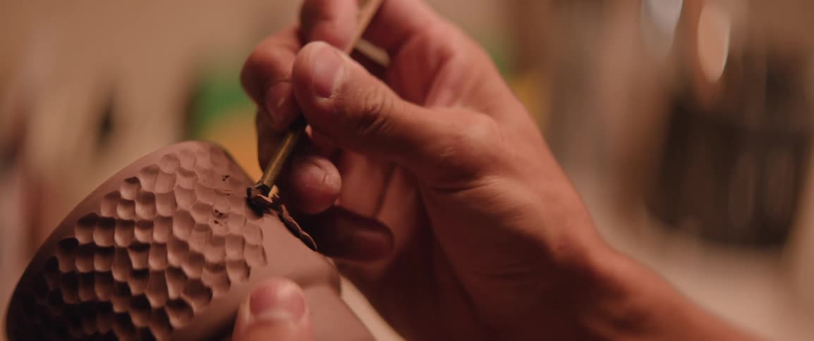 A person is intricately carving a textured pattern into a clay object using a small sculpting tool