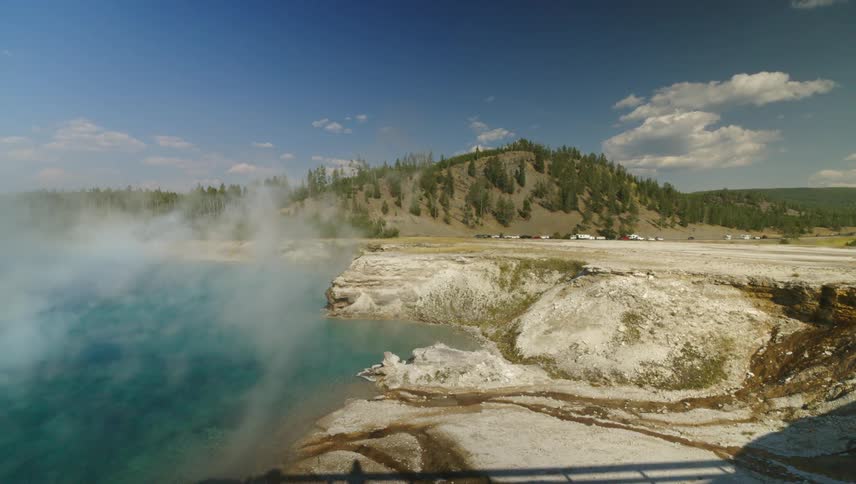 Steam rises form the hot spring as cars drive in the distance