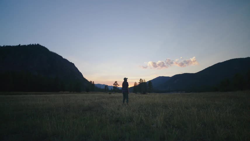 A woman wearing a cap is standing and looking sideways in a grass field with mountains in the background
