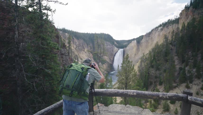 A person with a backpack taking pictures of a landscape with a waterfall between mountains