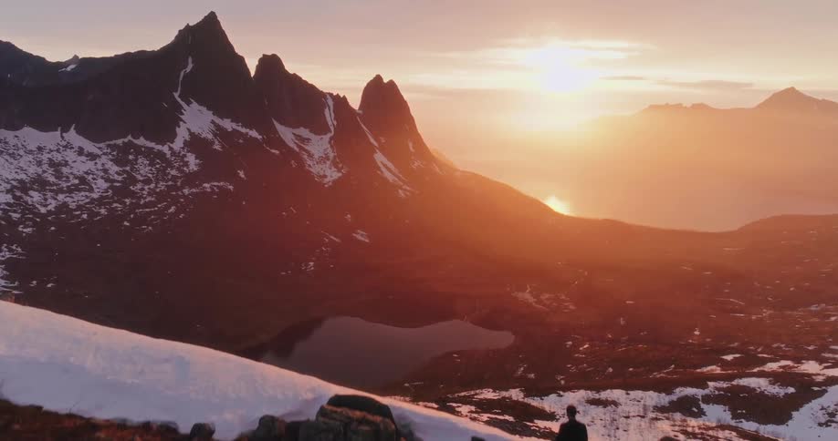 A person walks towards a rocky ridge at sunset overlooking jagged snowcapped mountains and a glowing horizon
