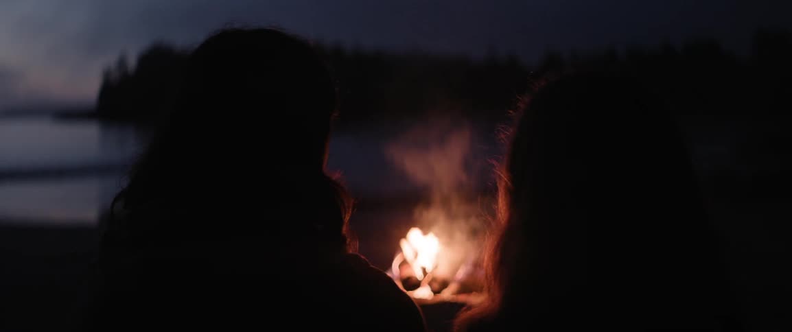 Two people sit on the beach looking at a small camp fire at night