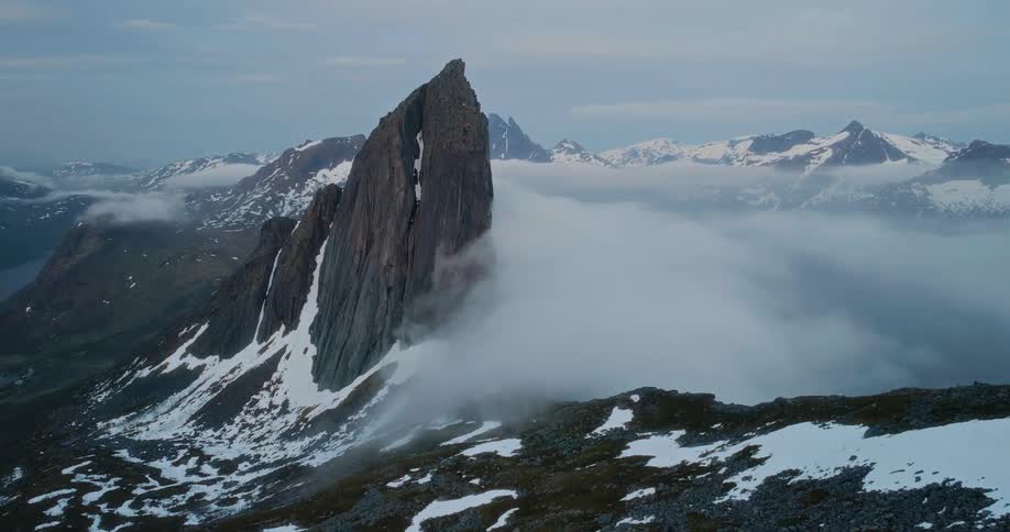 A dramatic jagged mountain peak rises sharply from a snowcovered landscape surrounded by mist and distant snowcapped mountains