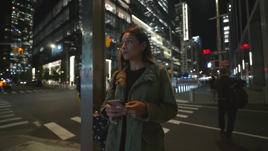 A woman is standing on the corner of a busy city street at night texting on her phone