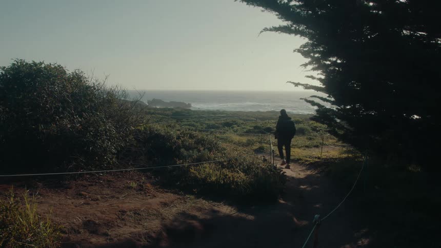 A male hiker walks along a trail near an ocean coast on a sunny day