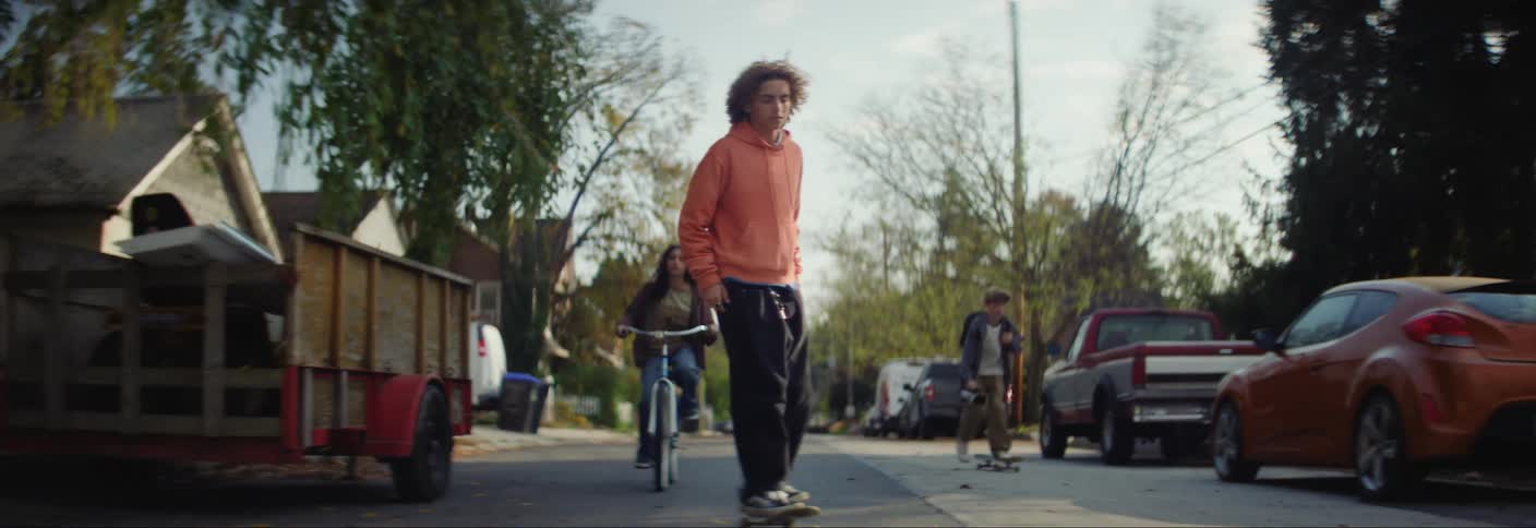 Three teenagers bike and skateboard down a residential street lined with fall trees