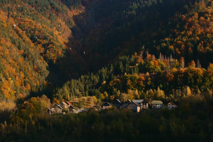 A small village is nestled in a valley surrounded by dense colorful autumn foliage on the slopes of a mountainous landscape
