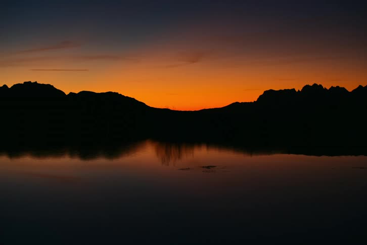 A landscape of a lake with still water reflecting dark mountains on a morning with orange sky