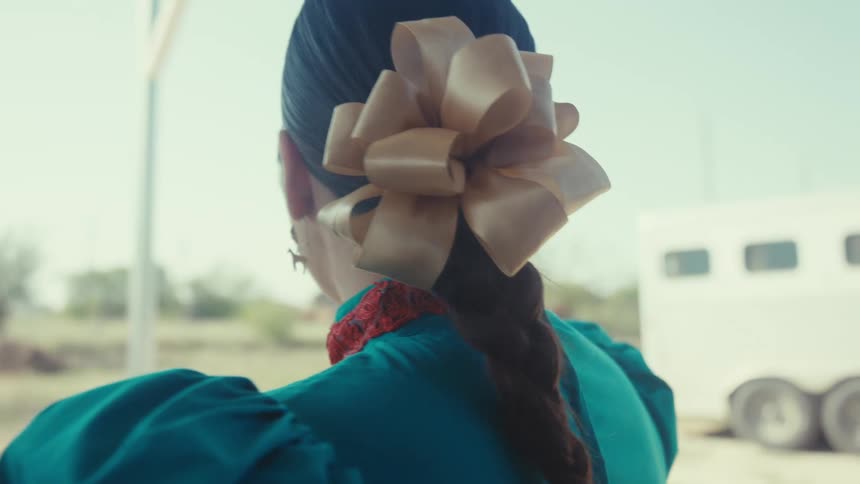 A woman with a long braid adorned with a large beige bow is seen from behind wearing a traditional blue garment with a red collar