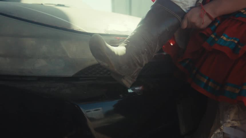 Closeup of a woman in a colorful dress adjusting her boot on the bumper of a car