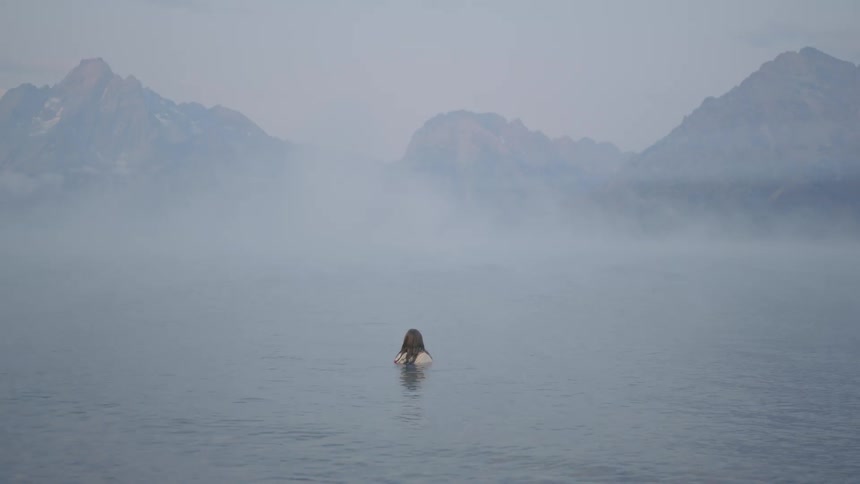 A woman submerges in a misty lake next to the mountains