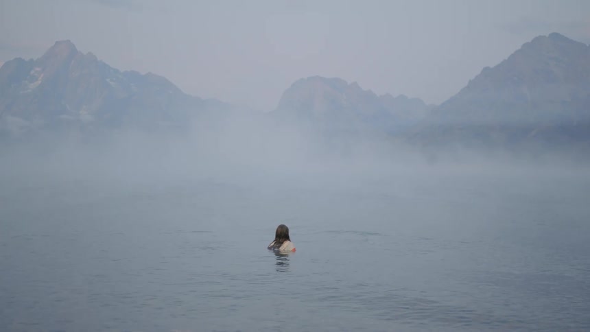 A woman fully clothed steps into a calm lake with misty mountains surrounding the scene