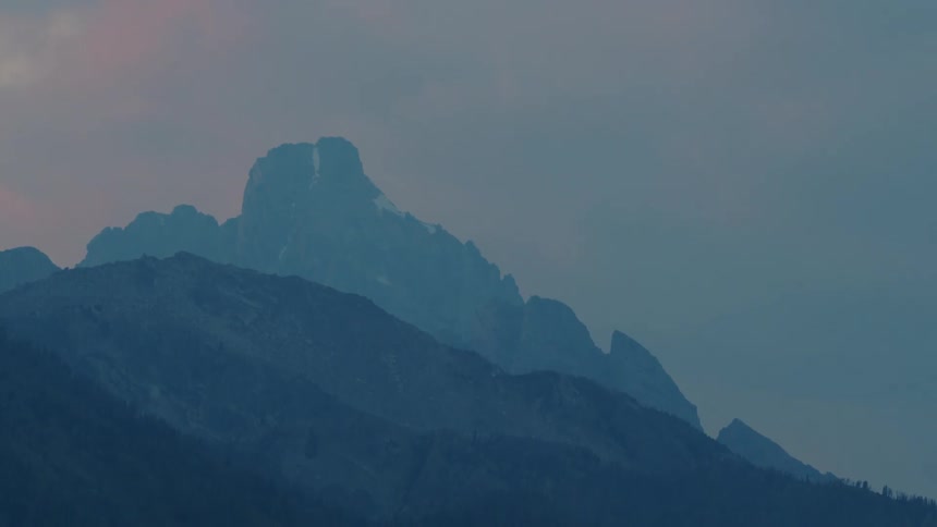 A serene mountain landscape with multiple layers of rocky peaks the tallest of which is partially covered in snow set against a cloudy sky