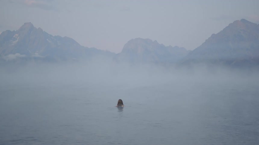 A woman floats on a calm lake surrounded by mist near the mountains