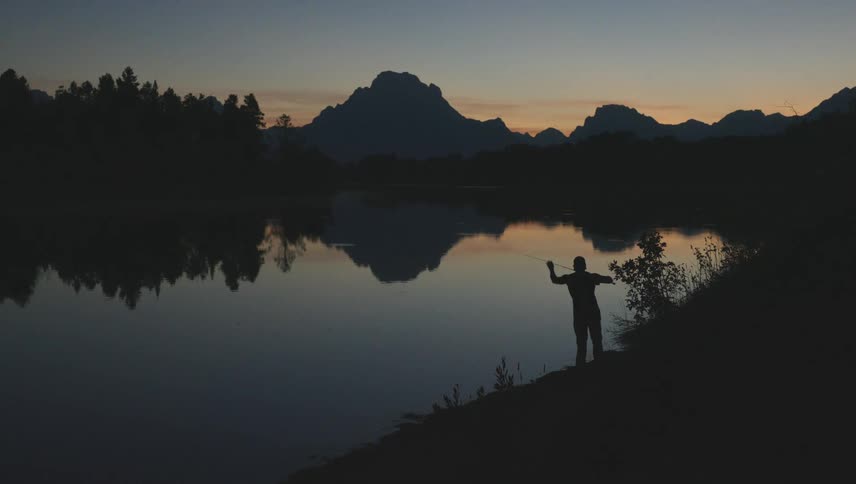 The silhouette of a person fishing in a lake with still water repeating the surrounding environment which is dark in the morning